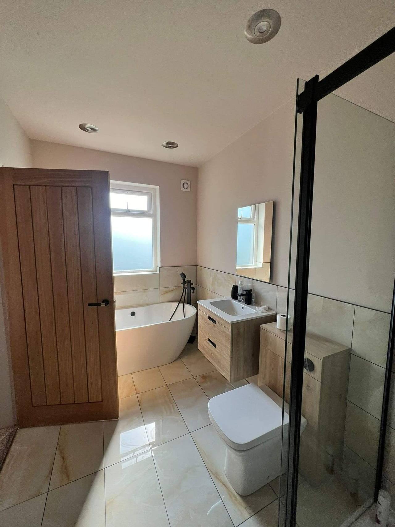 Renovated bathroom seen from the doorway: oak plank door, freestanding bath, wall-hung oak vanity and a black-framed shower screen